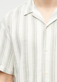 Λευκό πουκάμισο με κοντά μανίκια, κάθετες γκρι ρίγες και γιακά με χαραγή, κουμπωμένο από μπροστά, φορεμένο από ένα άτομο.