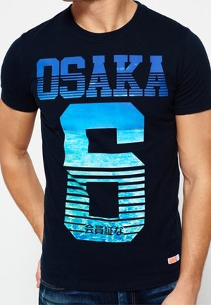 Mężczyzna w czarnej koszulce z napisem "OSAKA 6" w niebieskim gradiencie i wzorem morskich fal, oraz małymi japońskimi znakami pod numerem.