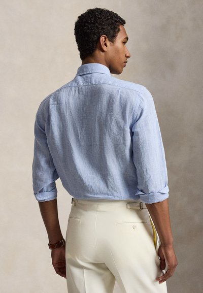 Homme portant une chemise bleu clair à manches longues retroussées et un pantalon couleur crème, vu de dos sur un fond uni.