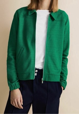 Person wearing a green zip-up jacket over a white shirt and dark high-waisted pants with silver buttons, standing against a plain background.