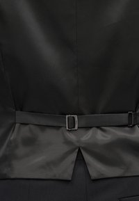 Rückseite einer schwarzen Anzugsweste mit verstellbarem Stoffgurt und kleiner Metallschnalle, die die Stoffstruktur und die mittlere Naht zeigt.