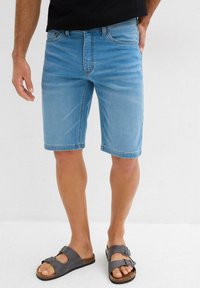 Shorts en denim bleu clair, longueur genou, avec un design classique à cinq poches. Portés avec des sandales grises à boucles réglables.