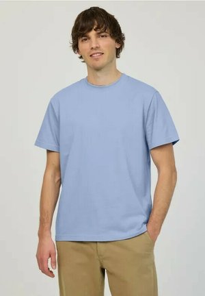 Ung mand stående med hænder afslappede, iført en lyseblå T-shirt med korte ærmer og beige bukser mod en ensfarvet baggrund.