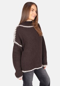 Brauner gestrickter Pullover mit hohem Kragen und weißen Akzenten an Schultern und Saum. Das Design ist übergroß und hat eine strukturierte Oberfläche.