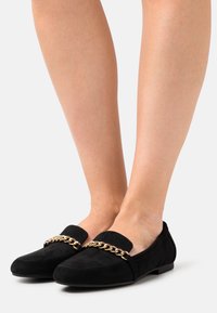 Černé semišové mokasíny s plochou podrážkou, zdobené zlatým řetízkem přes přední část a zaoblenou špičkou.