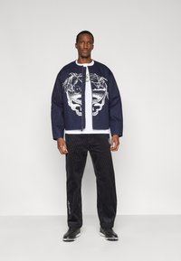 Veste en jean bleu avec un motif graphique, T-shirt blanc, pantalon en velours côtelé noir et chaussures noires. La veste est ouverte à l'avant et ornée de détails graphiques.