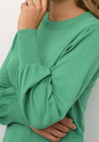 Personne portant un pull vert à manches longues avec des poignets côtelés, un bras croisé et l'autre main touchant leur visage près de la bouche.