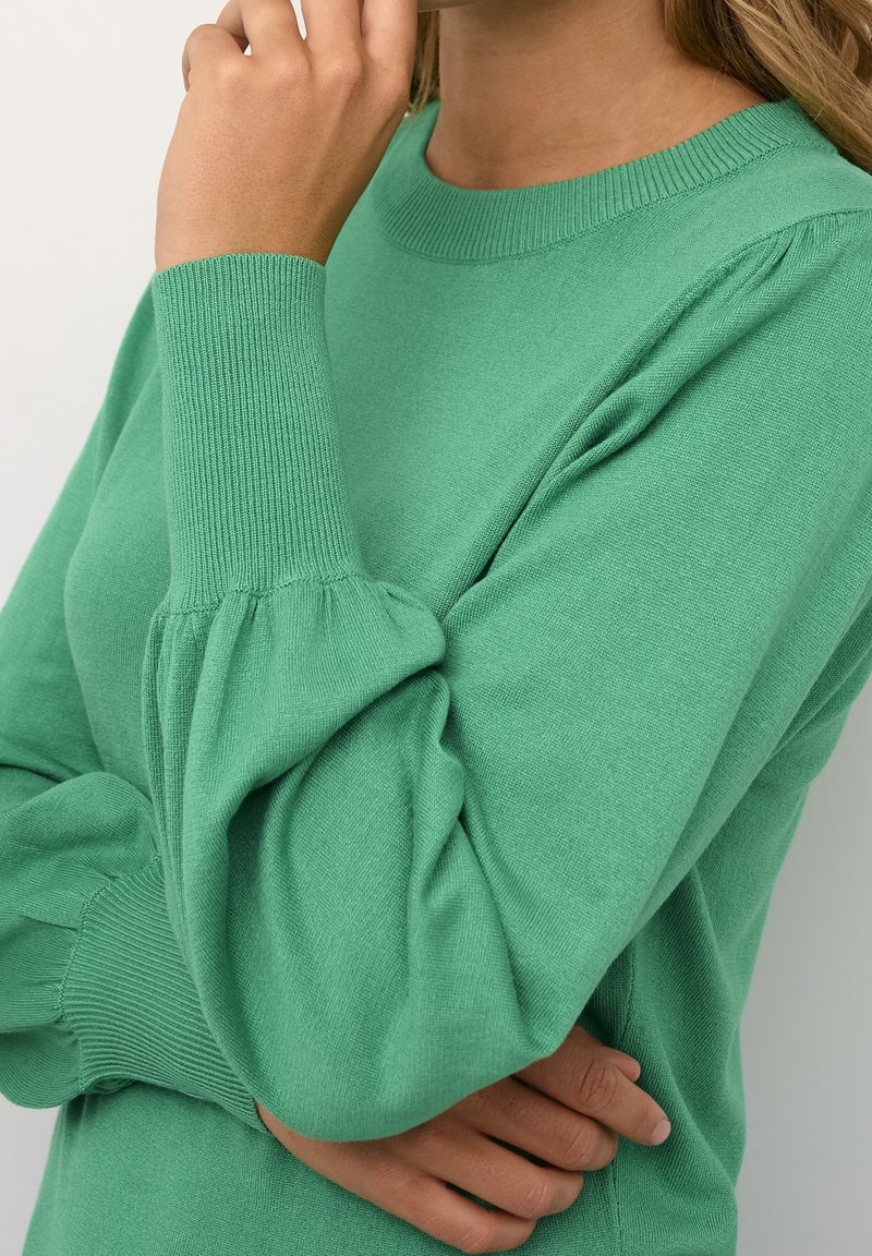 Personne portant un pull vert à manches longues avec des poignets côtelés, un bras croisé et l'autre main touchant leur visage près de la bouche.