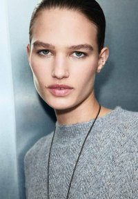 Suéter gris de punto con un patrón texturizado, combinado con un collar delgado negro. La piel suave, el cabello peinado y un maquillaje ligero realzan el aspecto.