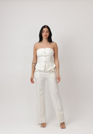 Vrouw met donker haar, gekleed in een strapless wit aansluitend korset met knopen en bijpassende witte wijde broek, staand tegen een effen achtergrond.