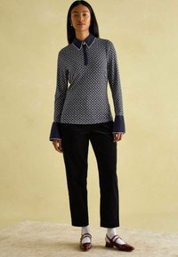 Navy-blau gemusterte langärmlige Polo-Shirt mit Kragen, kombiniert mit schwarzen Cordhose und burgunderroten Schuhen. Weiße Socken sind an den Knöcheln sichtbar.