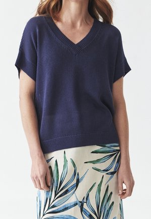 Kobieta ubrana w granatowy sweter z krótkim rękawem i dekoltem w serek, w połączeniu z białą spódnicą ozdobioną niebiesko-zielonymi motywami liści.