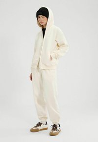 Crèmekleurige sweatshirt met capuchon en ritsluiting, gecombineerd met bijpassende joggers en sport schoenen met bruine accenten. Zwarte beanie.