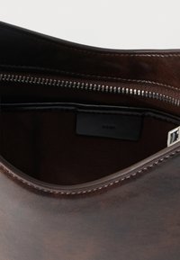 Καφέ δερμάτινη τσάντα μερικώς ανοιχτή που αποκαλύπτει ασημένιο φερμουάρ και εσωτερικό από σκούρο ύφασμα με μικρή μαύρη ετικέτα μέσα.