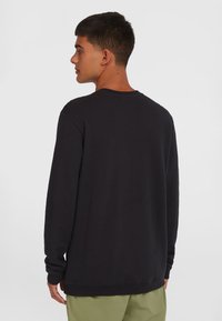 Maglione in cotone nero con maniche lunghe, collo girocollo a costine e texture uniforme, indossato da una persona di profilo.