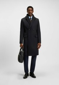 Manteau en laine croisé de couleur bleu marine foncé, avec un large col, deux poches avant et des boutons. Le modèle tient un sac noir texturé.