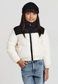 Weiß-schwarze Steppjacke mit hohem Kragen, Frontreißverschluss und elastischen Bündchen, kombiniert mit einer gerippten schwarzen Mütze. Jeans aus Denim sichtbar.