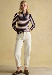 Blonda sieviete stāv ar rokām aiz muguras, ģērbusies tumši zilos svītrainos garos piedurkņu krekla, baltās sarullētās džinsās un brūnās loaferu apavās uz beidzīšu grīdas.