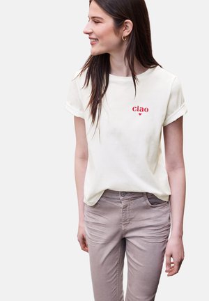 Jonge vrouw met lang bruin haar die lacht, draagt een wit t-shirt met opgerolde mouwen met "ciao" en een rood hart erop gedrukt, en taupekleurige broek.