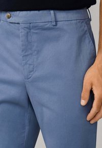 Vaaleansiniset puuvillapantalonit, joissa on sileä pinta, yhdellä napilla suljettava ja sivutaskut. Muotoiltu istuvuus ja siistit linjat.