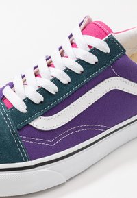 Gros plan sur une chaussure de sport violet et bleu sarcelle avec des lacets blancs, une bande latérale blanche, une languette rose et une semelle en caoutchouc blanche.