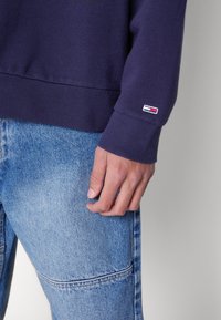 Donkerblauwe sweatshirt met geribbelde boorden en zoom, met een klein geborduurd logoplaatje. Gecombineerd met lichtblauwe spijkerbroek.