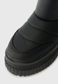 Černé kotníkové boty s hladkým, polstrovaným svrškem a texturovanou, vroubkovanou podrážkou. Mají zaoblenou špičku a viditelné švy kolem švů.