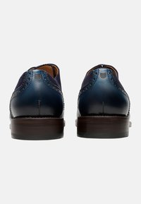 Scarpe in pelle blu navy con una texture liscia e dettagli brogue, dotate di un tacco in legno marrone chiaro e accenti blu scuro a contrasto.