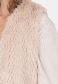 Chaleco de piel sintética rosa claro con superficie texturizada, sobre una camiseta crema suave y acanalada. Vista de cerca que resalta los materiales.