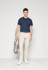 Navy t-shirt with pocket, cream-colored pants, brown belt, and white sneakers. Model holds a plaid jacket over his left arm.