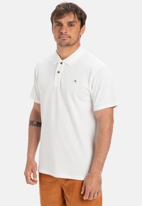 Biele polo tričko s klasickým golierom, dvoma hnedými gombíkmi a logom na pravej hrudi. Mäkká tkanina s krátkymi rukávmi.