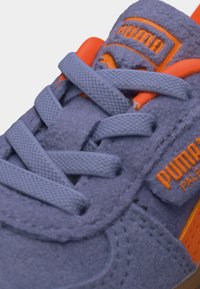 Κοντινό πλάνο μοβ suede αθλητικού παπουτσιού Puma με πορτοκαλί λογότυπο, κορδόνια και επένδυση στη γλώσσα.