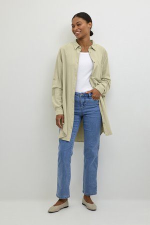 Donna sorridente in piedi che indossa una camicia lunga beige chiaro con bottoni, top bianco, jeans blu e ballerine beige su sfondo chiaro uniforme.