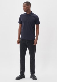 Námořnicky modrá polokošeľa s klasickým golierom a tromi gombíkmi, v kombinácii s čiernymi džínmi a čiernymi mokasínami. Hladká tkanina s minimálnym brandingom.