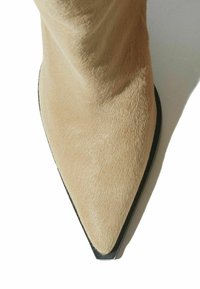 Botín de ante beige con tacón de aguja, punta afilada y textura suave, que presenta una suela plana negra y un diseño minimalista.