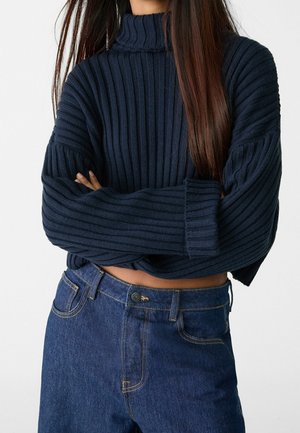 Femme aux longs cheveux portant un pull à col roulé côtelé bleu marine et un jean taille haute bleu foncé, bras croisés devant un fond uni.