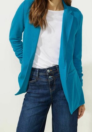 Teal blazer lavet af glat stof med en skræddersyet pasform, parret med mørkeblå jeans, der har kontrasterende knapdetaljer og forlommer.