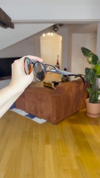 Ochelari de soare maro și negri cu lentile rotunde, având un cadru subțire și un mic accent portocaliu, ținuți în fața unui fundal neclar.