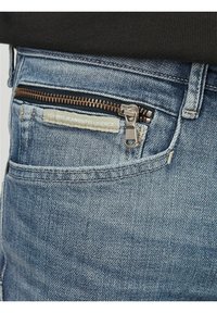 Jean en denim bleu avec une finition texturée, comportant une poche zippée avec des accessoires en bronze et une étiquette en tissu sur la taille.