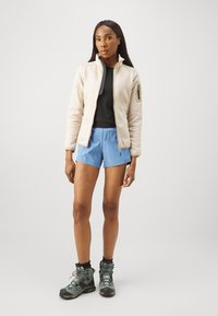 Fleecejakke i beige med glidelåslommer, svart topp, blå shorts og grå tursko. Glatt tekstur og skreddersydd passform.