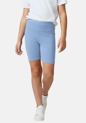 Kind draagt lichtblauwe knielange korte broek, wit T-shirt en witte sneakers, staand tegen een effen achtergrond.