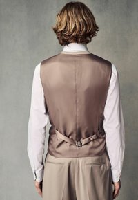 Gilet en satin marron avec une finition lisse, comportant une sangle dans le dos et une boucle en métal, porté sur une chemise blanche et un pantalon beige.