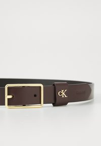Rudas odinis diržas su auksiniu stačiakampiu užsegimu, turi išraižytą "CK" logotipą ant laikiklio, lygus paviršius, klasikinis dizainas.