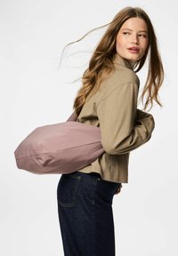 Bolsa rosa suave con forma redondeada y una correa larga, llevada sobre el hombro. La tela parece lisa, sin patrones visibles.