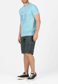 Helles blaues Baumwoll-T-Shirt mit einem Grafikdruck, kombiniert mit dunkelgrünen gemusterten Cargo-Shorts und dunkelblauen Sneakern mit beige Schnürsenkeln.