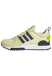 Light yellow athletic shoe with dark navy stripes, suede and mesh upper, white midsole, traction outsole, and a neon green heel accent.