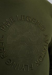 Sweat-shirt texturé vert olive avec du texte en relief et un motif en vagues. Le design inclut "PME LEGEND AVIATOR FLYING" affiché en évidence.