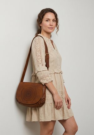 Femme en robe en dentelle beige portant un sac à bandoulière en cuir marron avec un motif circulaire en relief, debout contre un mur gris clair.