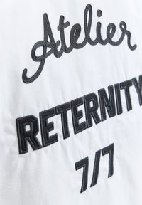 Texte noir brodé "Atelier RETERNITY 7/7" sur tissu blanc, mettant l'accent sur la typographie et la texture.