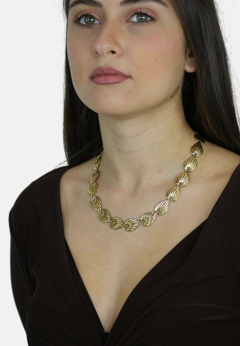Giovane donna con lunghi capelli scuri che indossa una collana dorata a forma di conchiglia e una maglia scura a V su uno sfondo neutro.